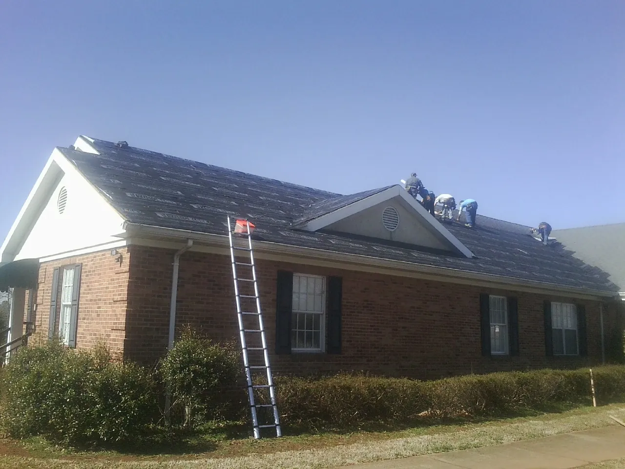 Crew tearing shingles off a brick ranch roof with a ladder set — Emergency Roof Repair work by our crew in Kauneonga Lake