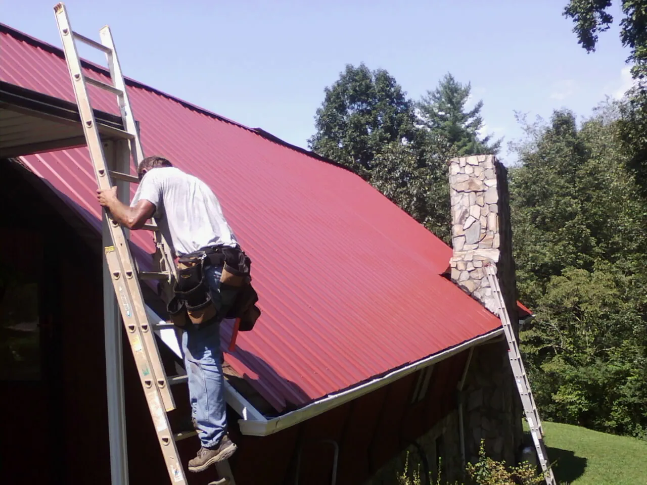 Roofer on a ladder beside a red metal roof and stone chimney — Soffit & Fascia Repair by Roofing Brothers in Kauneonga Lake